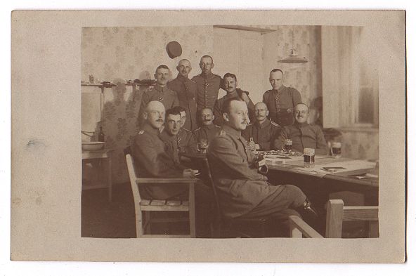 A group of German officers/NCO's? gather round one end of a long table (in the mess?), some standing, some seated.  There are some glasses of beer on the table.