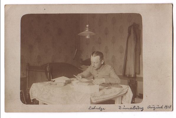 A man in uniform sits, reading, at a small, lace covered table which is covered in books and papers.  A single bed is in the corner of the room behind him, and a long coat hangs on a hook on the wall.