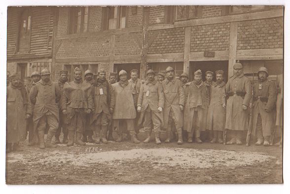 A large group of men wearing an assortment of uniforms pose in a muddy street in front of a timber framed building.
