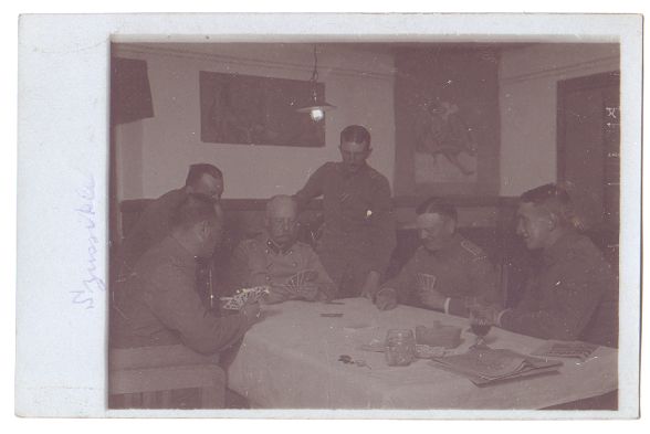 A small group of officers/NCO's sit round a table in the mess, playing a hand of cards.  A few others stand behind them, looking on.  A couple of glasses and a newspaper can be seen at the near end of the table.