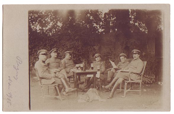 A group of officers or NCO's sit round a small table on a lawn, against a backdrop of bushes and shrubbery.  Some are reading, and there a few glasses on the table.