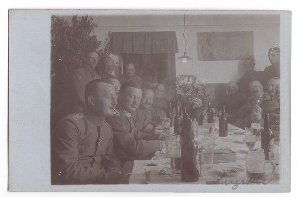 Many officers/NCO's are seated and standing around a table covered in bottles and glasses.  A box (cigars? cigarettes?) sits in the middle of the table, and a floral decoration can be seen at the far end.