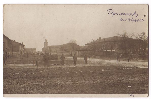 A view of a wide unpaved street or square, with men standing around in the distance. Several are standing near what may be a wooden water trough.  May be a scene from a prisoner of war camp?