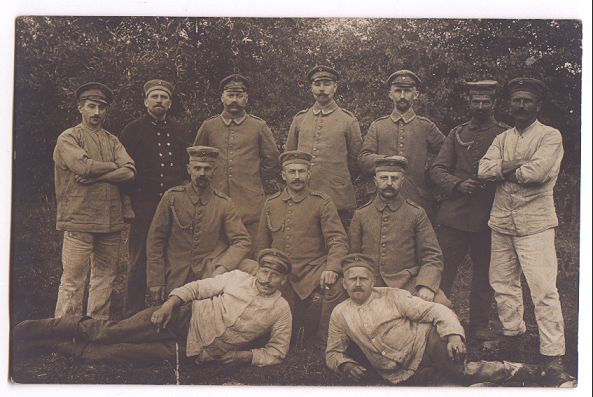 Twelve soldiers pose for a formal group photograph, in front of a tall hedgerow.  Seven men stand at the back, three are kneeling just in front of them, and two men are reclining on the ground at the front of the group.