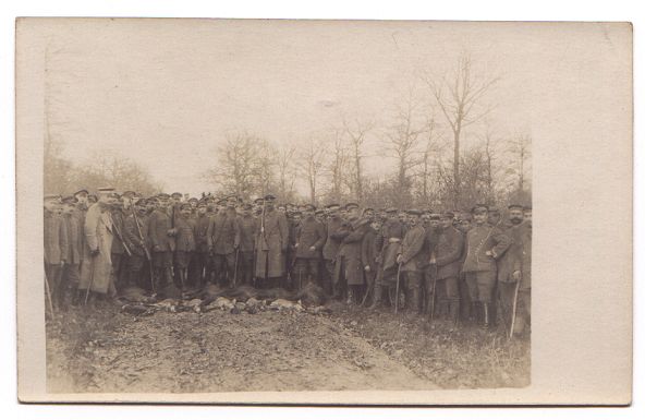 A large crowd of men, most wearing uniform, stand on a country track (with trees in the background).  They appear to have been hunting, and an array of animal carcasses are arranged on the ground in front of the men.