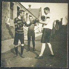 Two lads wearing knee-length shorts and boxing gloves and grinning, square up to each other. Possibly at training base?