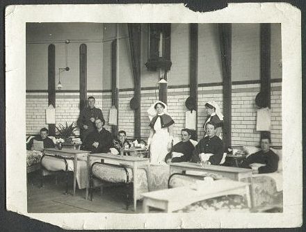 View of a neat row of hospital beds occupied by injured servicemen, with two nurses in attendance.
