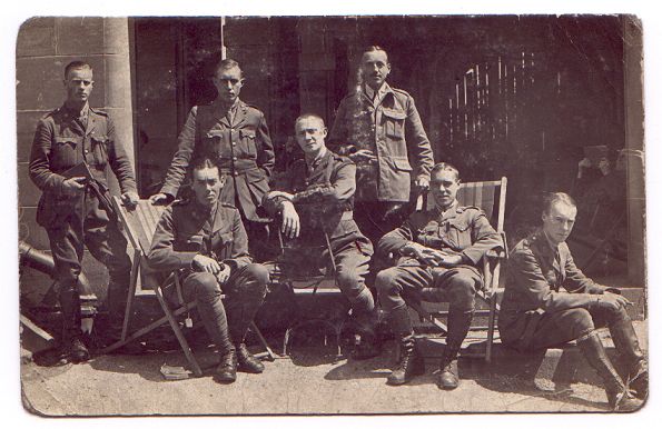 Seven officers pose informally. Three stand at the back left, three sit on a variety of chairs in the centre, and one sits on a step on the right.