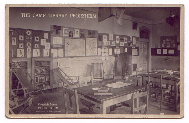 The room is filled with an assortment of tables and chairs, and the wall has many drawings and maps pinned neatly to it. A few shelves of books can be seen on the left of the picture.