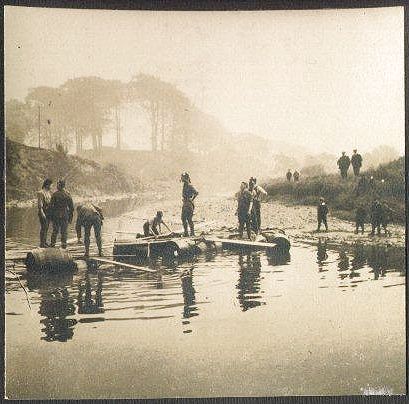 A misty river with some trees in the background.  Several men stand on the bank, and some in the river, assessing how to bridge the river using a collection of planks and empty oil drums.
