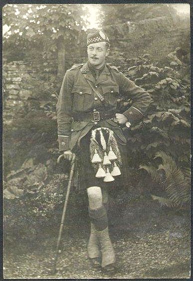 An informal portrait of Leslie, looking relaxed and at ease.  He is leaning lightly on a walking stick, and standing on a gravel path with fern covered boulders sloping steeply up behind him, topped by a rough stone wall.