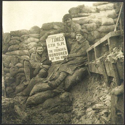Three soldiers, perched on a pile of sandbags in a corner of a muddy, sandbag walled trench, hold up a copy of the Evening Times showing the headline which fills the front page: '5th S.R. in the trenches: Honoured by the King'.