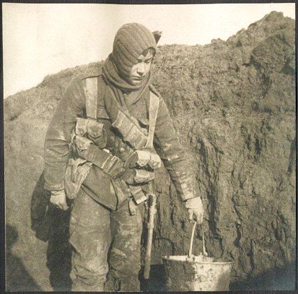 A young soldier stands in a muddy trench, carrying a bucket.