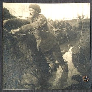 The soldier stands up to his knees in the water flooding the trench, wrestling with a tangle of barbed wire.